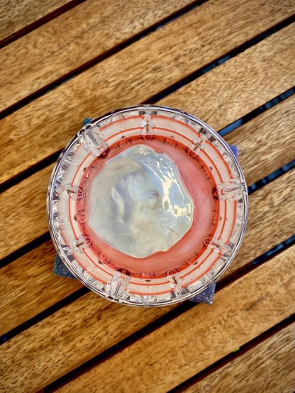 a top-down angle of a white cocktail with crystal clear hand-shaped large rock of ice and Peychauds bitters floated on top, sitting on a wooden table