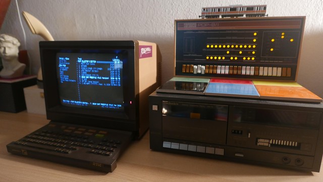 The image features three vintage electronic devices placed on a wooden surface against a white wall. On the left, there is a Minitel 1, with a screen displaying blue-white text, and a keyboard with colorful keys. On the right, there is a PiDP8/i computer with a digital display of blinkenlights. It is on top of a cassette deck.