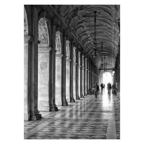 This is a black and white portrait format image of an ornate colonnade in St Mark's Square in Venice. Light falls through the columns from left to right, highlighting the tiled floor. The angles in the image form a focal point to the middle right, and people are silhouetted against the daylight at the end of the colonnade. Taken with a Sony RX100.