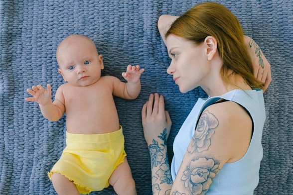 Ein Baby mit blauen Augen und hellgelbem "Windelröckchen" und eine an den Armen mit Blumenmustern tätowierte junge Frau mit rötlichem Haar und hellblauem, ärmellosem Oberteil liegen auf einer blauen Decke. Die Mutter ist dem Kind zugewandt.