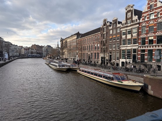 A view across a broad canal in central Amsterdam, with canal boats moored to the right, and a series of terrace buildings on view behind them.