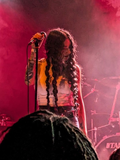 A singer on a stage. She in the center of the frame, all bathed in a red and warm light. She is holding a microphone, which lays on its stand, with her right hand while she is looking to the right of the picture, away from the microphone. She was long curly dark hair and dressed a white top. Tatoos are visibile in the arm that is holding the microphone.