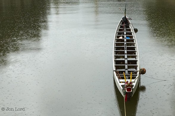 A Simple Low Contrast Colour Photo In Landscape Format. Nearly All The Image Is Filled With The Still Waters Of A River. Neither Bank Is In View. The Central Area Stretching From The Top To The Bottom Has A Wide Bright Strip Of Reflected Light From The Overcast Sky. The Left And Right Edges Have A Dark Green Tint. The Entire Surface Of The River Has Round Splash Marks From Falling Rain. To The Right Of Centre And Aligned With The Camera Is A Long Dragon Boat. It Is Moored Bow Facing The Camera And Only A Short Distance Away. Forward Of The Bow Is The Bowsprit In The Form Of A Colourful Dragon, Jaws And Eyes Wide Open. Aft Are Fourteen Positions For Her Crew With A Rowlock On The Sten For A Paddle/Tiller In Order To Steer. 
The Location: The River Silver In Mui Wo, Hong Kong (2016).
