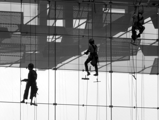 Three window cleaners in harnesses are suspended on ropes, working on the exterior glass facade of a building.

Drei Fensterputzer sind an Seilen aufgehaengt und arbeiten an der aeusseren Glasfassade eines Gebaeudes.