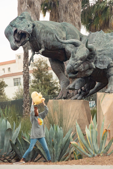Photo of Lety and the Creature Mastodon stuffed toy beneath the sculpture of a T-Rex and Triceratops in combat outdoors in Los Angeles during the day. From top to bottom, Lety is seen wearing a gray graphic sweater featuring a rainbow seal of the Kennedy Space Center. She's standing in the dirt in front of some agaves, holding the creature above her head while they look up and backward towards the elevated “Dueling Dinosaurs” sculpture by Douglas Van Howd.