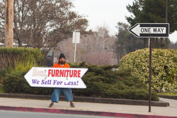 next best thing: photograph of a street with a ONE WAY sign. A man on the sidewalk is carrying a large arrow-shaped sign that says "Best FURNITURE We Sell For Less!"