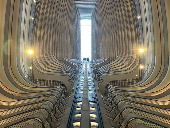 Looking upwards along an elevator shaft. The “train” in the center is the elevator track. The light in the distance is the skylight. The curves are various layers of balconies.