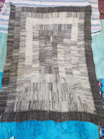 A hand knit afghan-sized blanket is spread out to dry on some beach towels. The blanket is a rectangle made in a spiral, starting with a strip in the middle, which turns each corner at right angles. The colors shift organically from creamy white to dark charcoal gray. Running down the center of each strip is a line of small lace motifs resembling a cat track.