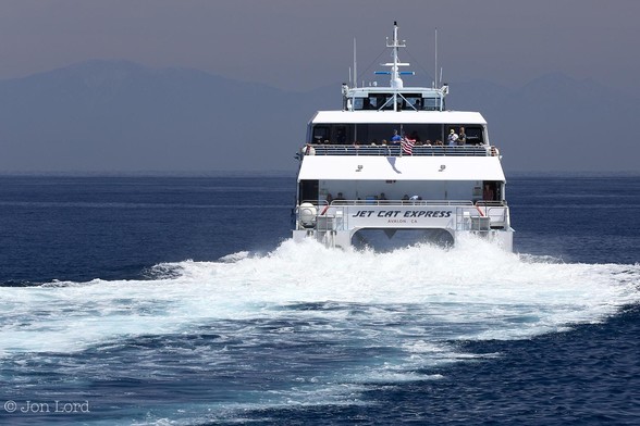 This Is A Colour Seascape Photo In Landscape Format. Covering The Lower Two Thirds Is An Expanse Of Dark Blue Calm Sea. To The Right Of Centre Is The Stern Of A SeaCat Fast Ferry Under Way.  The View Of The SeaCat Is Aligned So None Of The Sides Are In View, Just The Stern. Aft Of The Ferry Is A Mix Of Blue Ocean And White Froth From The Powerful Water Jet Nozzles. The Ship Being A Catamaran Has Two Parallel Hulls Separated By Five Or So Metres With Two Main Decks And A Bridge Above The Upper Deck. The Aft Section Of Both Decks Are Open And Passengers Can Be Seen. Some Distance Ahead Of The Ferry Is A Tall Mountainous Ridge That Stretches From The Left To The Right Margins. On The Left Side A Little Late Spring Snow Can Just Be Made Out. Above The Sky Has A Slight And Very Light Orange Hazy Tint To It. 
The Location: Santa Catalina, California. The Body Of Water Is The San Pedro Channel (The Pacific) And The Mountain Range Is The San Gabriel Mountains. The Ferry Is The 'Jet Cat Express' Bound For Long Beach (2015).
