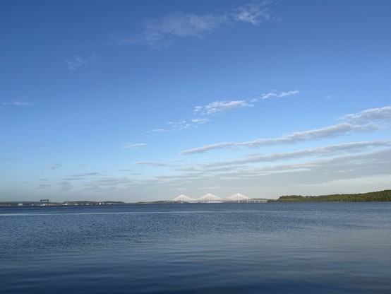 A serene view of a calm body of water under a blue sky with scattered clouds. In the distance, a bridge with a distinctive design spans across the water, flanked by green landscapes on either side.