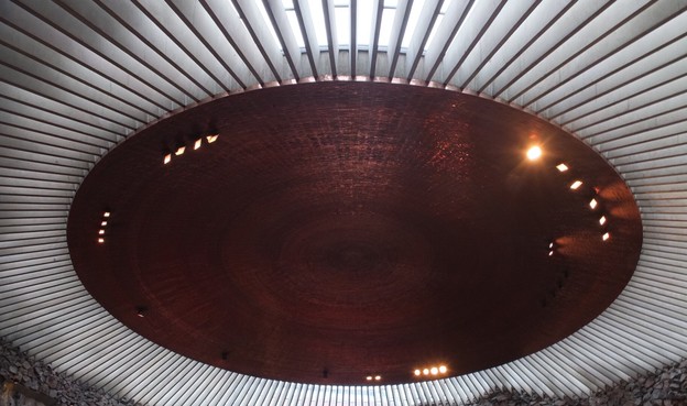 This image captures the interior of a circular ceiling design. The ceiling features a smooth, dark surface with a wood-like texture, surrounded by a series of vertical beams or slats. Soft lighting is integrated into the design, highlighting the curvature and creating a warm ambiance. The overall composition emphasizes the geometrical circular structure.