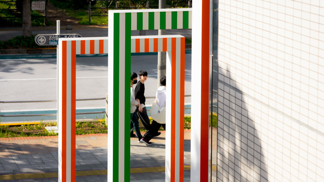 The image shows a set of vertical and horizontal structures with orange, white, and green stripes near a walkway. In the background, a couple of people are walking on the sidewalk alongside a street. The wall of a building with a tiled pattern is visible on the right side.