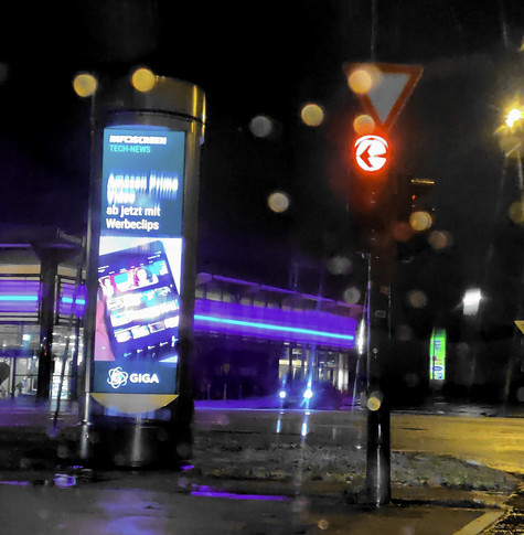 Nachts, direkt neben einer Ampel: eine über 4m hohe Display-Säule mit "Infoscreen Tech-News: Amazon Prime Video ab jetzt mit Werbeclips" - einem Bild dazu und "GIGA" als "Nachrichten"-Verbreiter. Es ist etwas Regen auf der Scheibe.