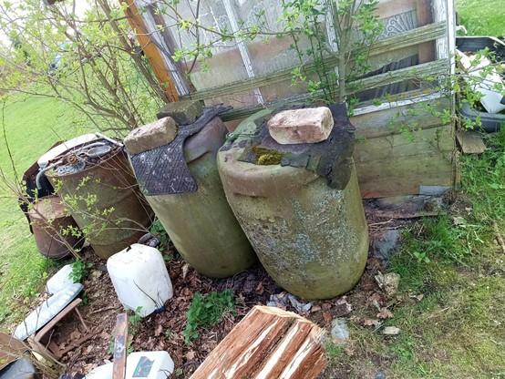 Two huge green plastic barrels with lichen growing on them hiding behind a small shack. They are covered with rubber mats and bricks. Behind is also a steel oil drum.