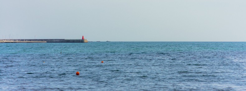 The image shows a calm sea with a few scattered orange buoys on the water. In the background, there's a breakwater with a red structure, possibly a lighthouse or marker, on top. The sky is clear and blends softly with the horizon.