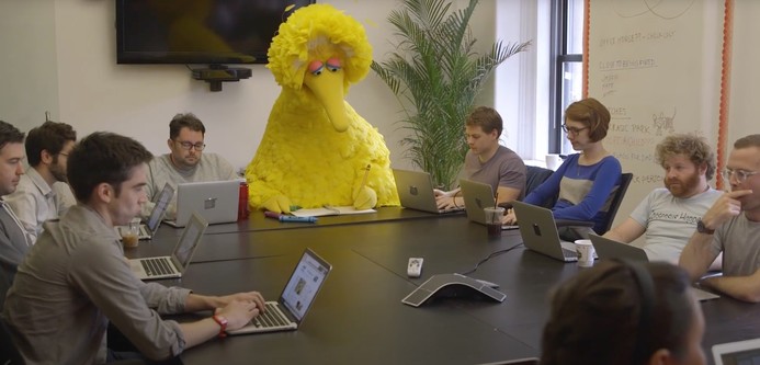 Big Bird (a Sesame Street character that is a large, yellow bird) sitting at one end of a conference table, drawing with crayons on a pad of paper, surrounded by a group of young human (not bird) comedy writers, all with laptops open in front of them.