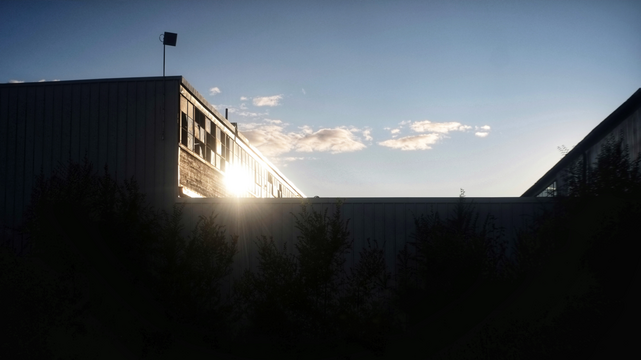 The top of an abandoned factory is hit by the setting sun causing a bright reflection off the windows. The blue sky has some thin clouds and the bottom half of the image is pure black.