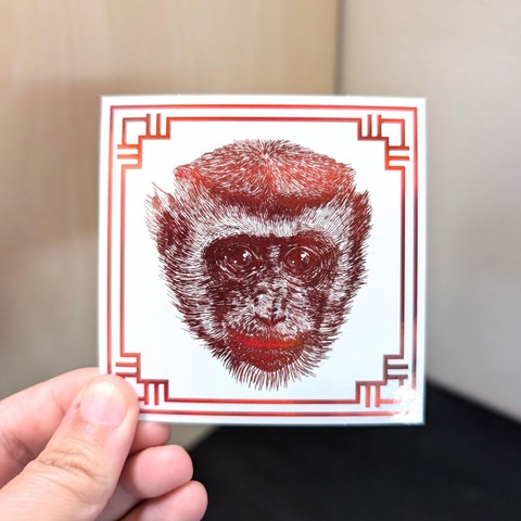 Photo of a left hand holding a square sticker. The sticker is a holographic drawing of the Chinese Zodiac animal: the Monkey.