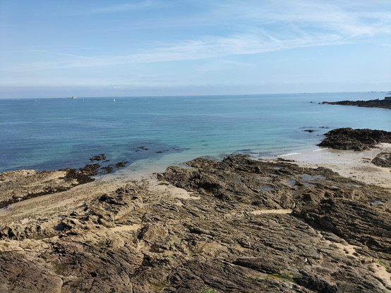 Vue depuis un promontoire rocheux sur une plage de sable à marée basse, bordée de formations rocheuses sombres. La mer est calme, d’un bleu turquoise, avec quelques voiliers à l’horizon sous un ciel bleu légèrement voilé.