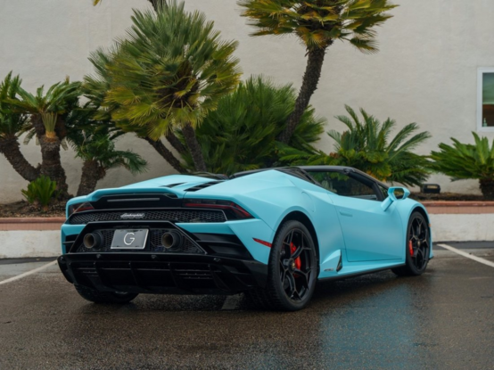 a photo of the vehicle seen from behind in a 3 quarter view, the convertible top down, it is in  a parking lot, shubs growing against the building in the background