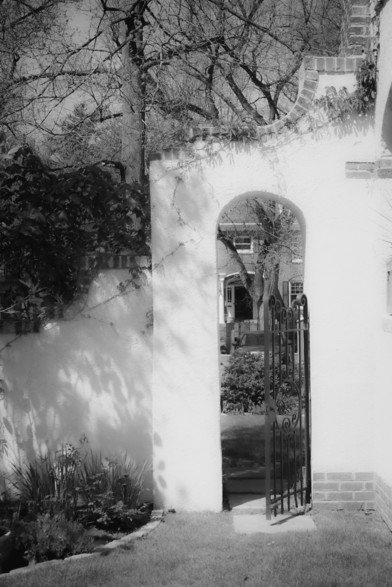 The image depicts a black and white scene featuring an arched gateway with a wrought iron gate. The gateway is part of a white wall with brick accents at the base. The gate is open, revealing a view of a garden and a house in the background. The garden includes various plants and shrubs, with a tree visible to the left of the arch. The house has a brick facade and a visible window. The scene is framed by bare tree branches, suggesting a winter or early spring setting. The sunlight casts shadows on the wall, adding depth to the image.

Provided by @altbot, generated privately and locally using Ovis2-8B