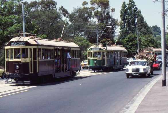 A Melbourne W1 class tram in brown and cream livery, displaying RACECOURSE on its desto and a supplementary board saying '1927 Vintage Tram' leads a Melbourne W2 class tram in green and cream livery with a blank desto and its own supplementary board displaying '1928 Vintage Tram' along a gently curving street with parkland in the background.  Cars are running parallel to the trams, most prominently at right a white Mercedes Benz sedan towing a trailer loaded high with what appears to be garden prunings.