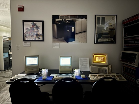 A Mac SE, Mac Plus and DECmate III on a table with various posters and artwork.