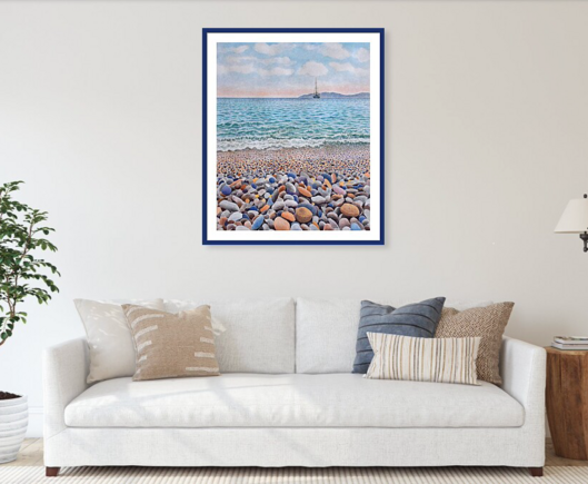 A large prints of a watercolor painting on a wall. A coastal landscape, showing a rocky beach and ocean waves. It hangs over a couch with pillows.