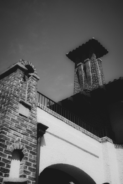 The image is a black and white photograph of a building with a prominent brick tower. The tower features a square base with a tiled roof and is constructed from dark bricks, with visible mortar lines. The building has a white stucco facade with a smooth texture, contrasting with the rough brickwork. An arched opening is visible at the base of the tower, and a small arched niche is present on the brick wall. A black metal railing runs along the top of the building, and a clear sky is visible in the background. The image is taken from a low angle, emphasizing the height of the tower and the architectural details.

Provided by @altbot, generated privately and locally using Ovis2-8B