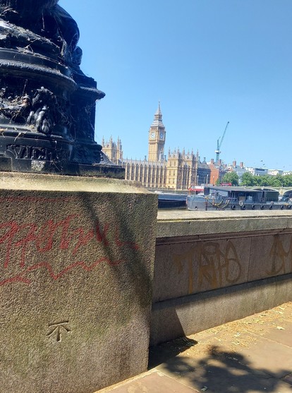An OS arrowhead benchmark in the stone, or concrete, wall of a walkway alongside the River Thames. The Palace of Westminster and "Big Ben" are in the background.