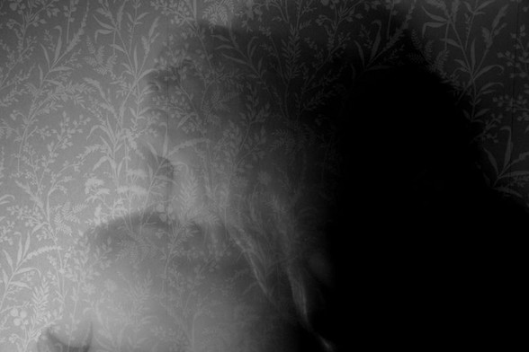 A black and white portrait of a woman's head and a little below her shoulders. She stands in front of an interior wall, seen obliquely from the side with her head tilted slightly back. It looks as if she is looking at something high up to the left. Her hair hangs down on the right side and so does her dark shadow. The pattern on the wallpaper behind her is filled with various printed grasses against a vaguely darker background and it shines through the woman, as if she were transparent.