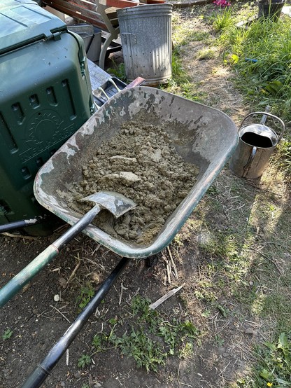 A wheelbarrow full of cement. There is a spade laid across the barrow. A watering can sits on the floor nearby.