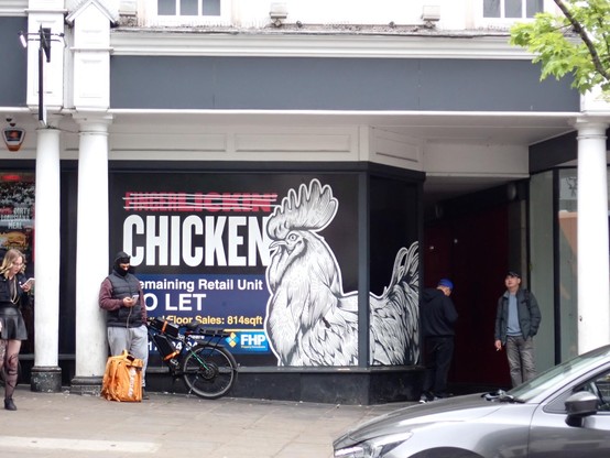 A large graphic image of a chicken fills the glass of a shop window. Above the phrase "Finher Lickin" has four white lines (tape perhaps) crossing it out. a blue "To Let" sign is propped up at the bottom. Of the window.