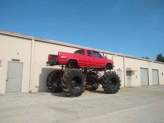 A ridiculously jacked up red pickup truck with giant tractor tires parked behind a beige building.