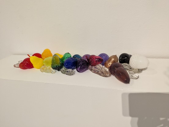 A bunch of acorns and seeds rendered slightly larger than life in a rainbow of bright glass colors on a white pedestal