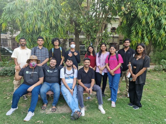 A group of 13 people - 4 women and 9 men - in a park, looking at the camera. 4 are sitting on a bench, and the others are standing. Two are wearing respirators, and two are wearing grey OpenStreetMap India T-shirts.