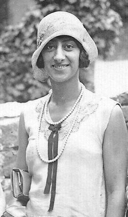 Black-and-white photo of a woman smiling, dressed in a 1920s style outfit with a cloche hat and a pearl necklace.