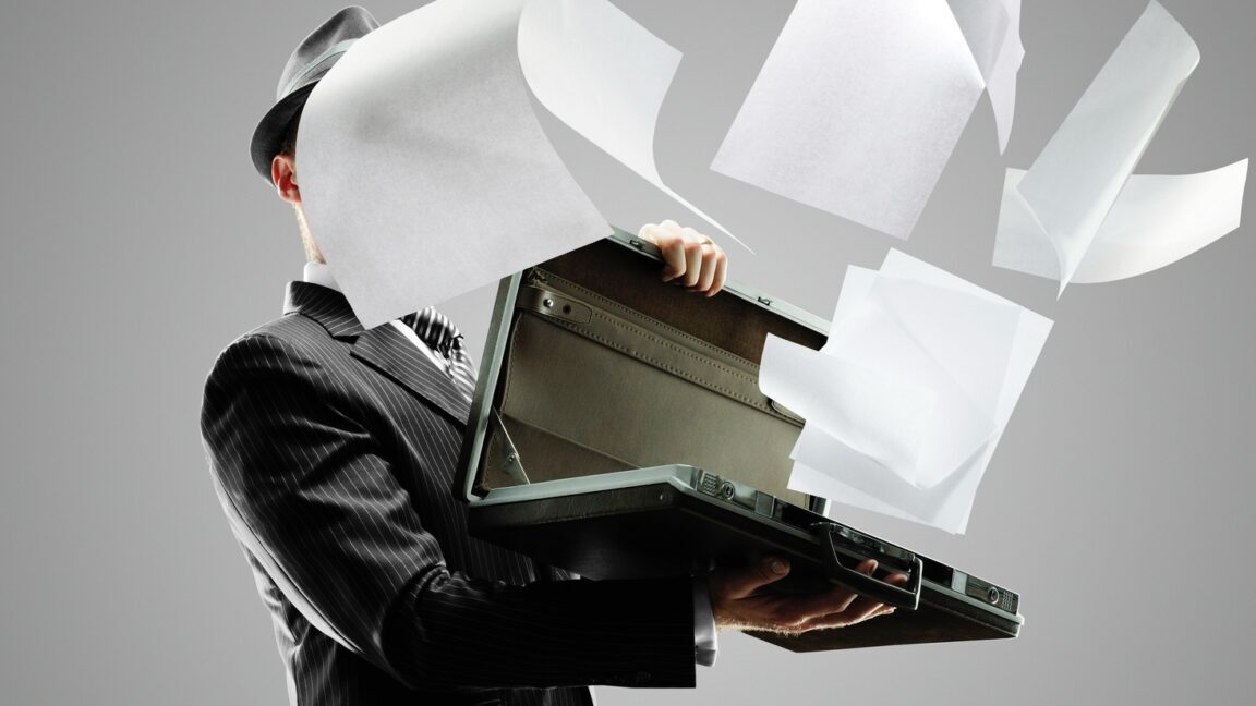 Man in dark pinstripe suit holding open a briefcase, from which papers are flying out and covering his face.
