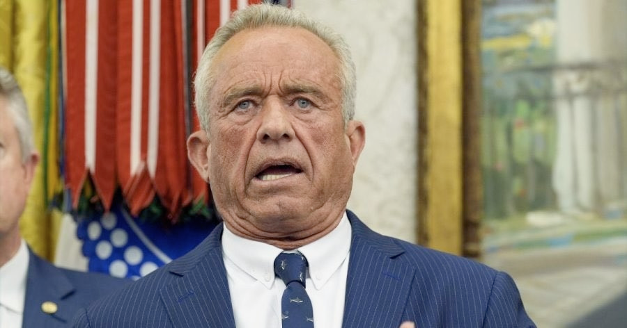 The image features an older man with grey hair, dressed in a formal suit and tie, speaking into a microphone. The background showcases an American flag and appears to be an indoor setting, possibly a formal event or press conference, with an ornate, official-looking interior. The man's expression appears serious, contributing to a formal atmosphere. The image behaves as a possible meme template, particularly for content involving serious, humorous, or ironic commentary.