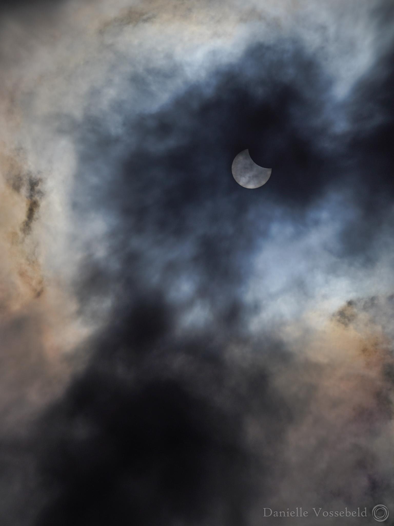 Partial solar eclipse surrounded by dark and white-ish clouds, with some colours in it. 