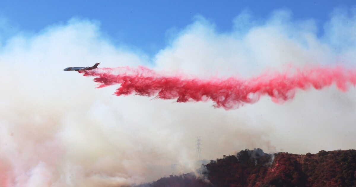 A long trail of red follows an airplane over hills.