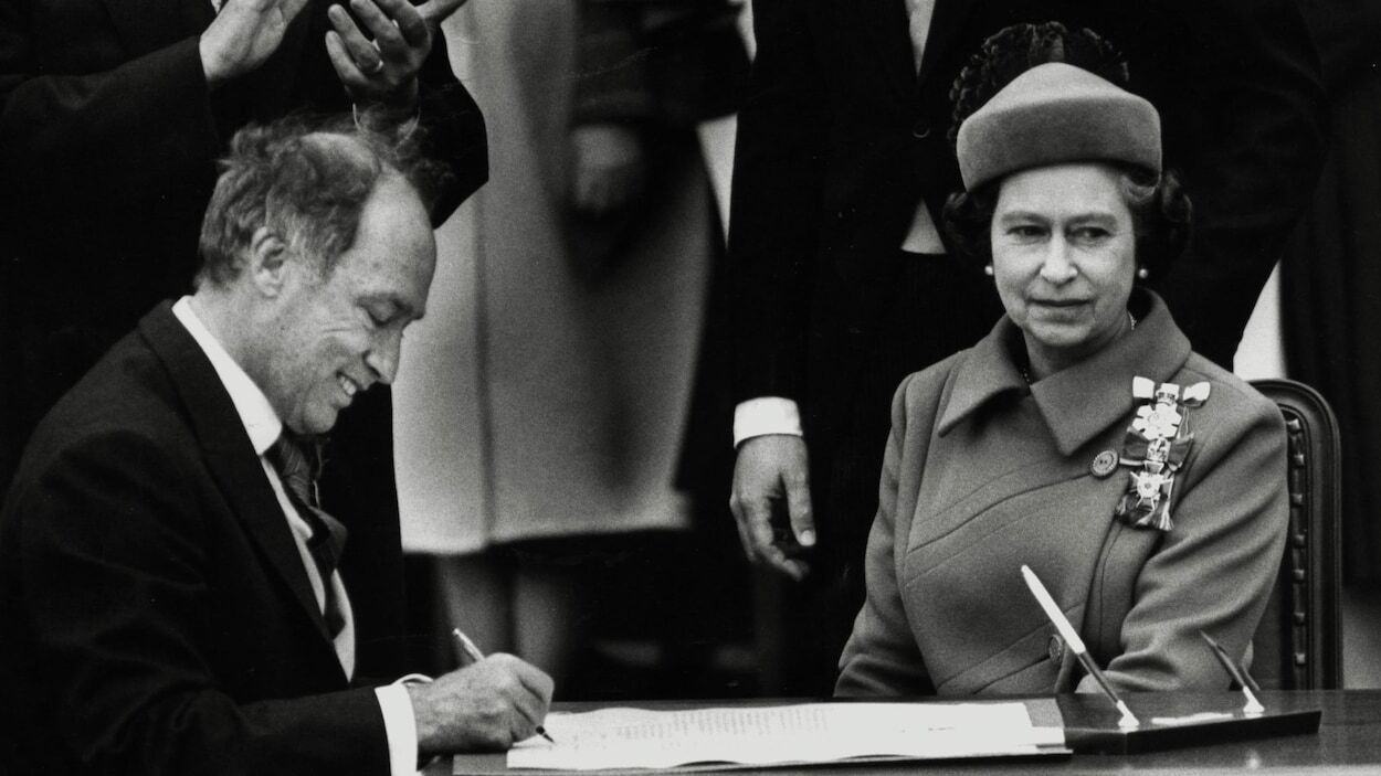 Pierre Elliott Trudeau sourit en signant un document devant la reine.