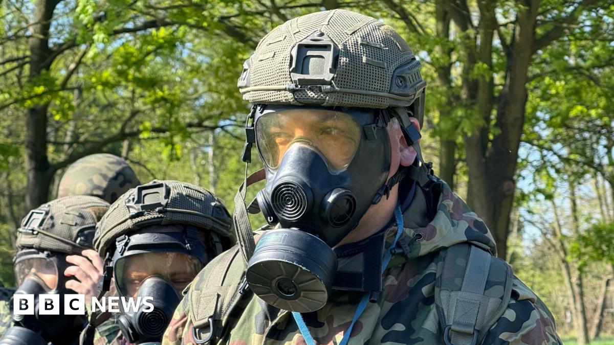 A close-up of a young man wearing a gas mask, helmet and army camos. Behind him are three women wearing the same. The group is outdoors in a sunny field with trees.