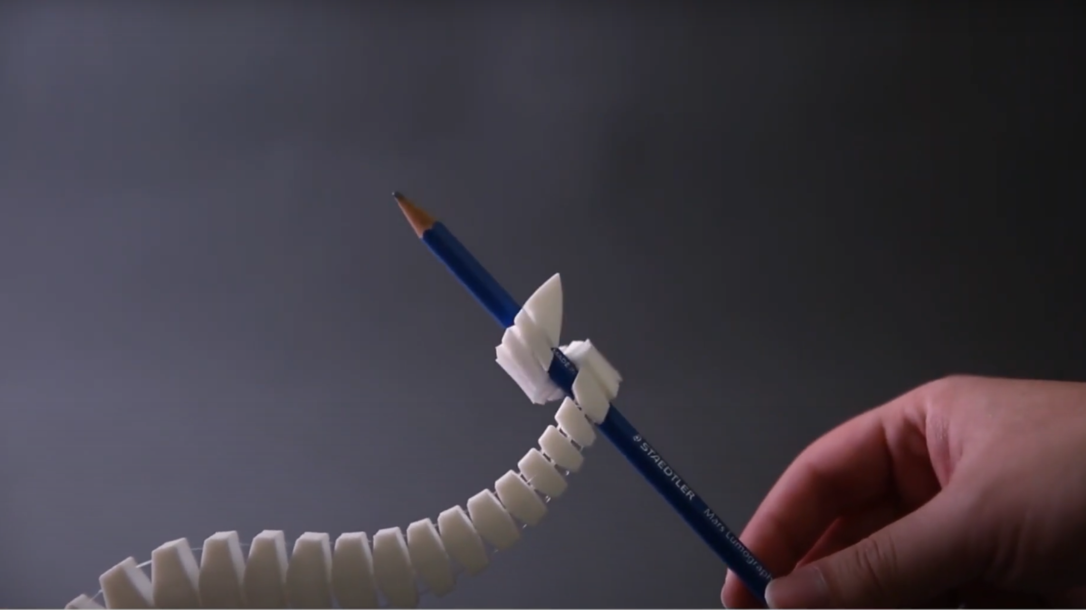 A human hand is shown in the bottom right corner of the picture, holding one end of a pencil. A white, segmented, mechanical tentacle extends from the bottom left corner of the image and wraps around the other end of the pencil. A human hand is shown in the bottom right corner of the picture, holding one end of a pencil. A white, segmented, mechanical tentacle extends from the bottom left corner of the image and wraps around the other end of the pencil.