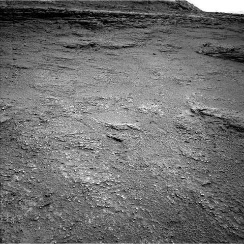 A grayscale photo from the Martian surface shows rough, slightly uneven, dark gray, rocky terrain extending to a plateau that rises in the distance, at the top of the frame. In the foreground, many smaller rocks are poking above the surface, much lighter-toned than the surrounding ground, looking like white specks on the surface.