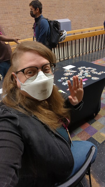 A blond woman sitting at a table covered in stickers and waving while taking a selfie 