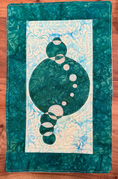 a small quilted piece of art with intersecting circles and a wave with alternating colors. It is made with batiks with a teal and light blue.