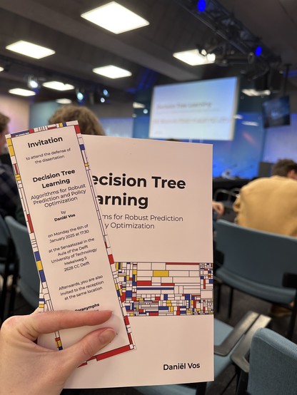 View from the audience of a conference room. I am holding up a dissertation and bookmark. Their designs are clearly inspired by the work of Piet Mondrian.

The bookmark contains the following text:

Invitation

to attend the defense of the dissertation

Decision Tree Learning

Algorithms for Robust Prediction and Policy Optimization

by
Daniël Vos

on Monday the 6th of
January 2025 at 17:30

at the Senaatszaal in the
Aula of the Delft
University of Technology
Mekelweg 5
2628 CC Delft

Afterwards, you are also
invited to the reception
at the same location