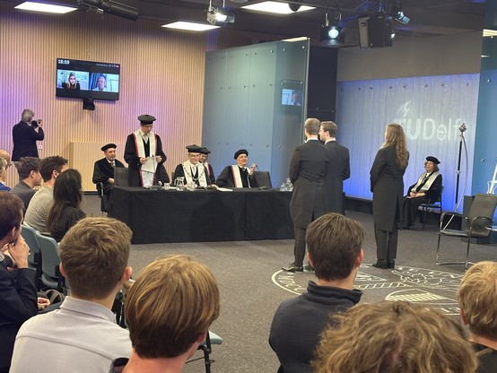 A lecture room, seen from a seat in the audience. There is a black table with five professors in black robes and hats. One of them is standing and reading from a piece of paper. Seen on their backs are three young people in black suits, standing in front of the professors, listening to the speech. In the background is a monitor with two people joining on Zoom, the Beadle sitting in a chair, also wearing black robes and a hat, and a photographer. The TU Delft logo is projected on the wall.