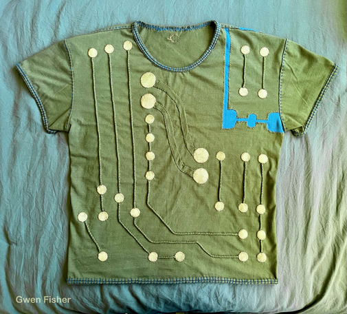 Photo of a t-shirt embroidered with a circuit design. The shoulder has a section added that looks like it’s a separate board that has been snapped apart along a line of perforation. The shirt is covered in gold circles that are appliquéd onto the shirt. The circles are connected with lines of couching, couched strips of fabric. Everything is hand sewn. The neck line and all hems are finished with the same double line appliqué stitch treatment.
